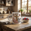 Faith-themed ceramic mug with pink roses, butterflies, and gold cross, on a wooden table with plants and sunlight